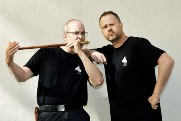To menn i svarte skjorter. Han til venstre spiller fløyte.