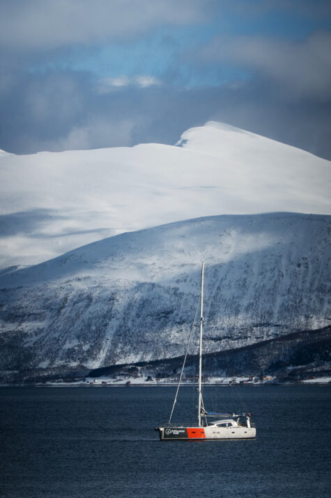 Hvit og rød båt på mørkt hav med snøkledde fjell i bakgrunnen.