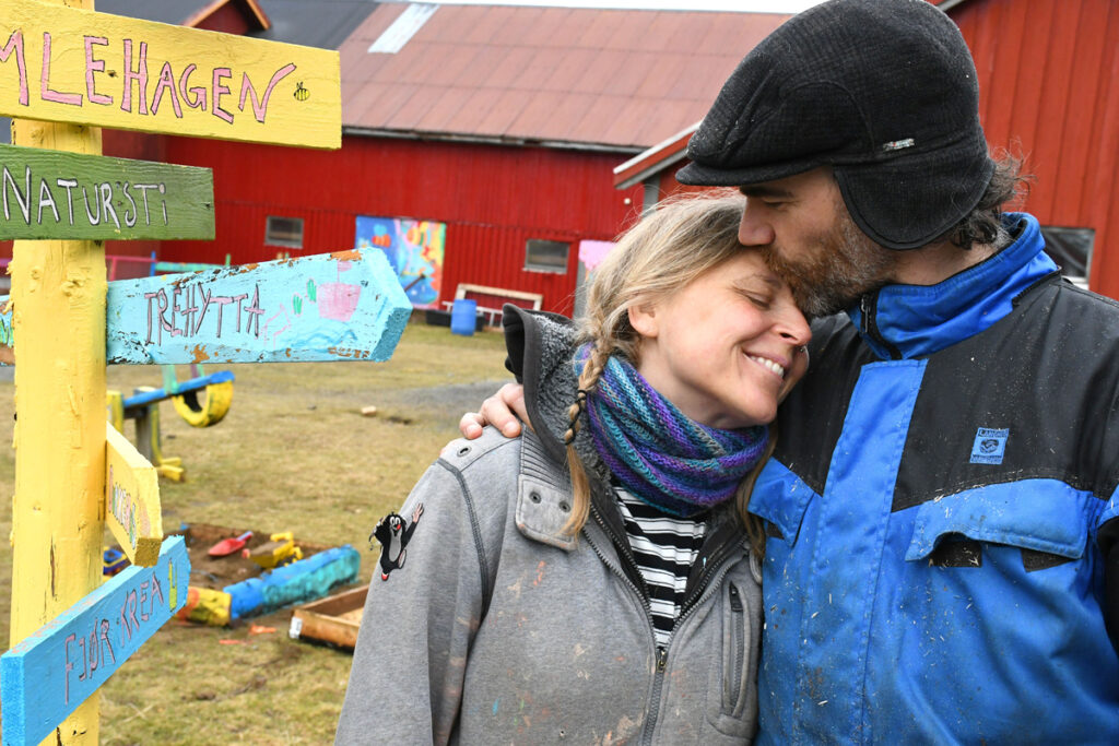 To personer står tett sammen og klemmer hverandre. Rød gårdbygning i bakgrunnen.