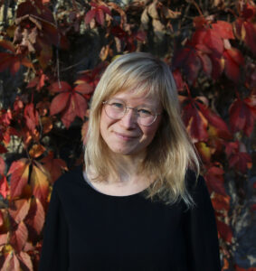 Person med lyst hår i svart genser foran løvverk i høstfarger.