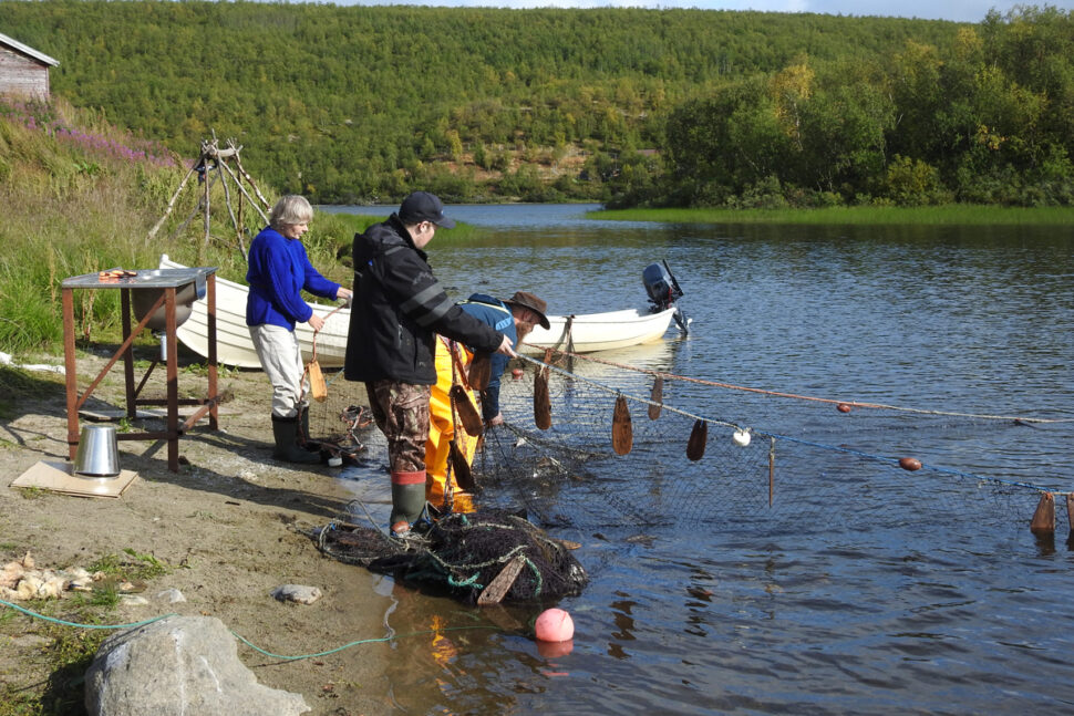Mennesker båt og not i vannkant med grønt landskap i bakgrunnen.